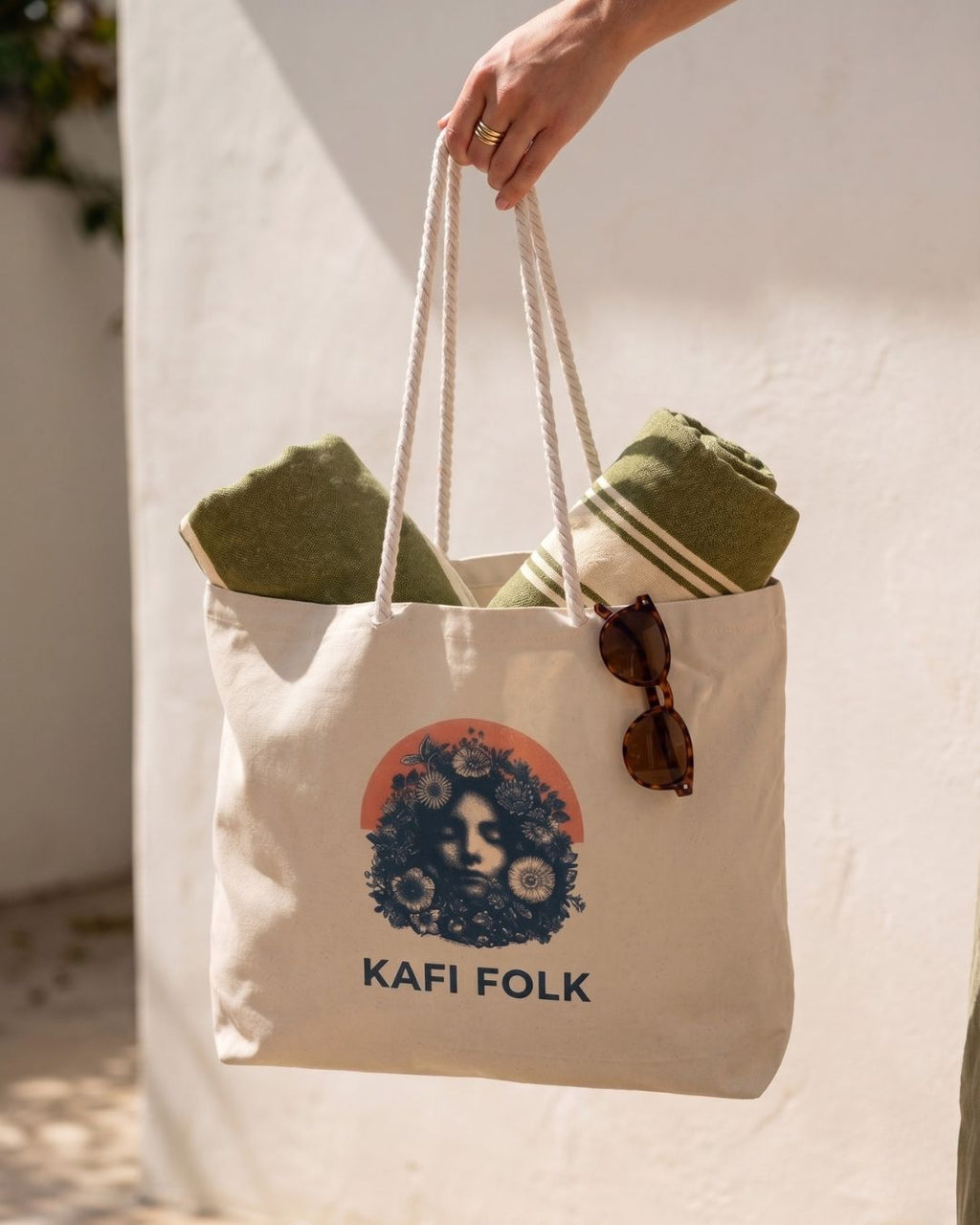 Person holding a tote bag with 'Kafi Folk' design, containing green fabric and sunglasses.
